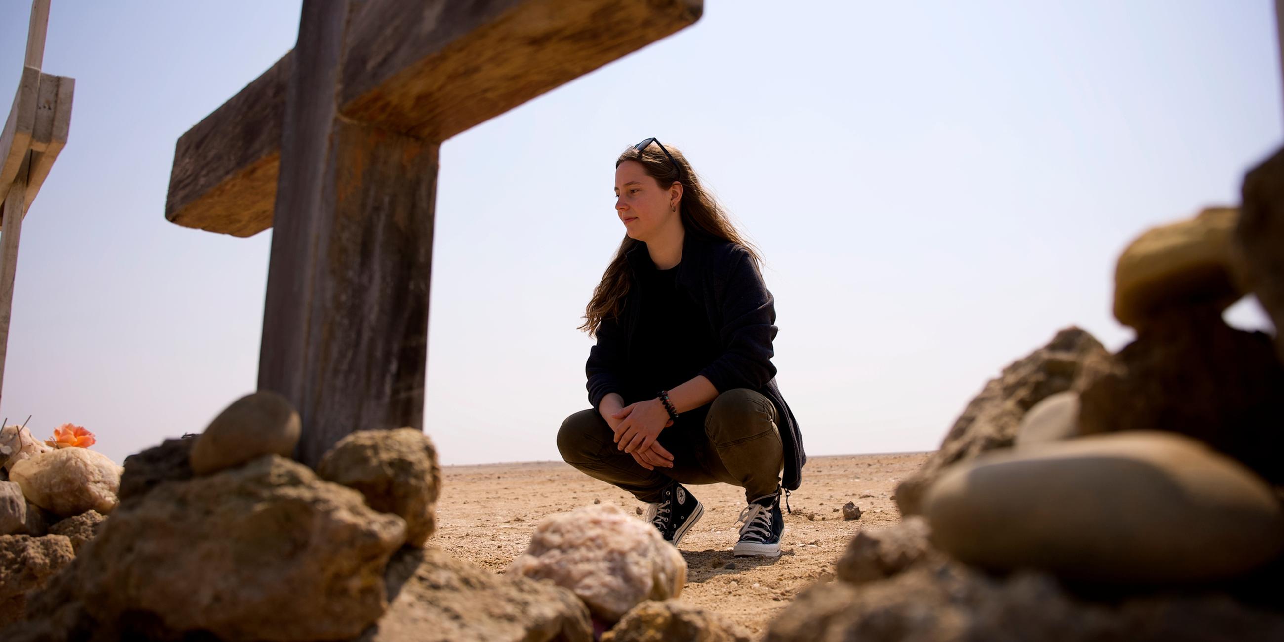 Antonia Joschkos hockt vor einem Kreuz in der Namibischen Wüste und schaut dieses an. Ein weiteres Kreuz steht mittig im Vordergrund, am rechten Bildrand ist ein weiteres Kreuz im Anschnitt zu sehen.
