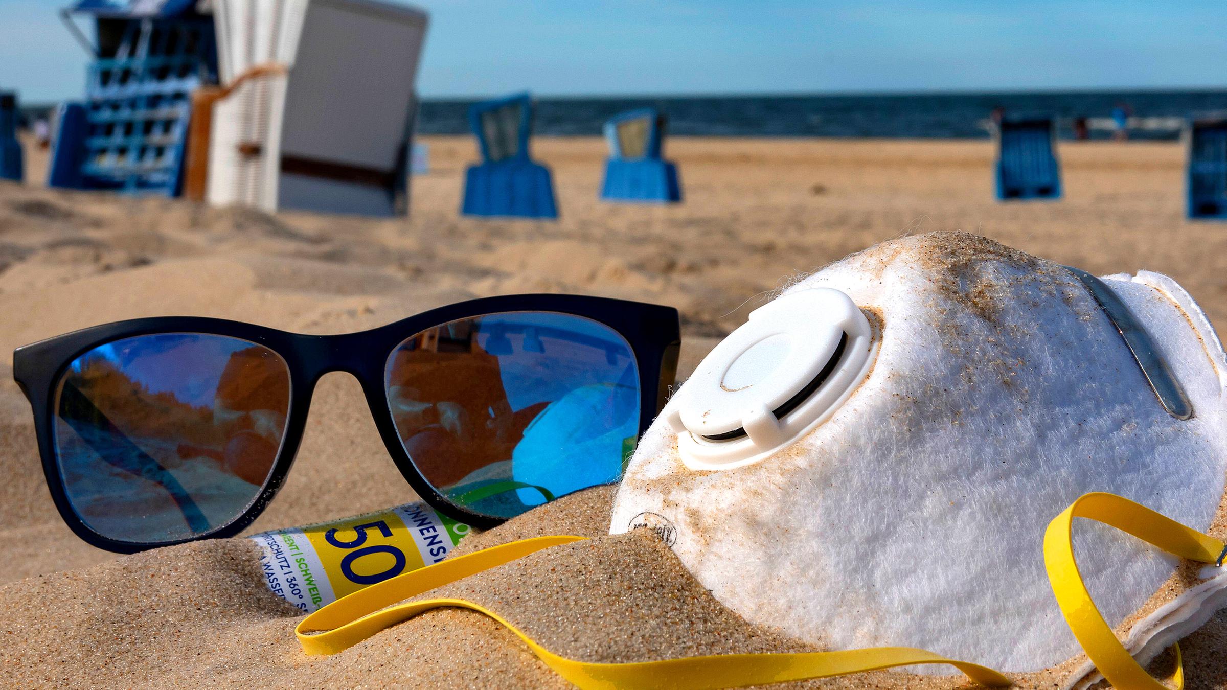 Ein Mund-Nasen-Schutz liegt neben einer Sonnenbrille und einem Sonnenspray im Sand.