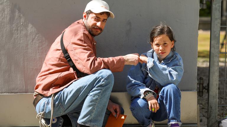 "Ready.Daddy.Go!": Michel (Fridolin Sandmeyer) und Getsimani (Fee Schuster-Böckler) sind vor einer Mauer in der Hocke und halten ihre Fäuste aneinander. Sie gucken zuversichtlich nach vorne. Michel hält einen orangefarbenen Umschlag in der Hand.