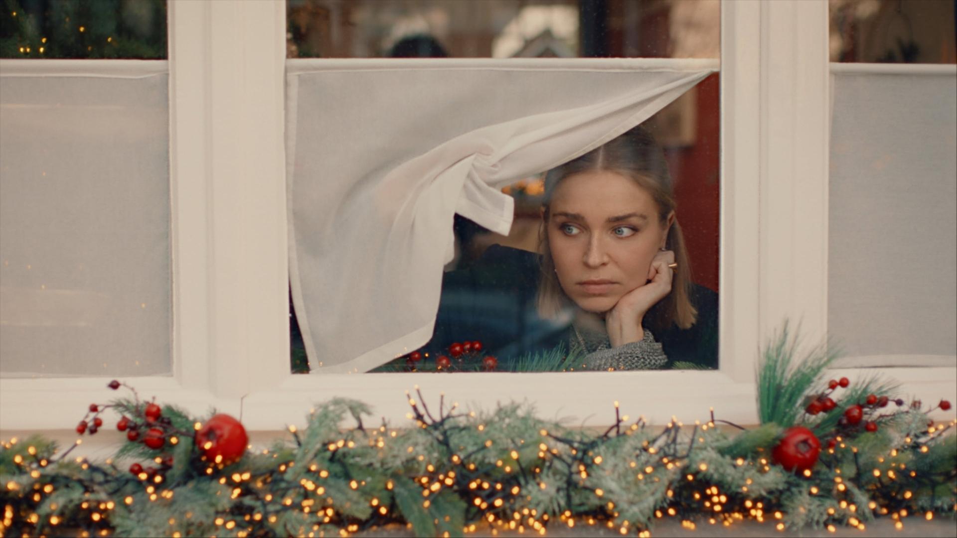 "Single Bells - 12 Dates till Christmas: Alex": Mina (Julie Van den Steen) blickt prüfend aus einem Fenster auf die Straße. Die halbhohe Gardine hebt sie mit einer Hand zur Seite. In die andere Hand stützt sie ihren Kopf. Auf dem Fenstersims außen liegt leuchtender Weihnachtsschmuck.