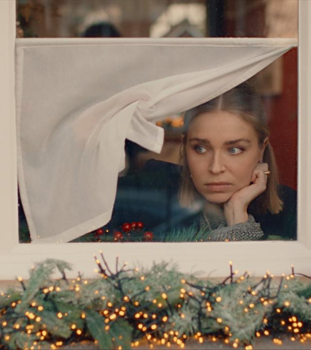"Single Bells - 12 Dates till Christmas: Alex": Mina (Julie Van den Steen) blickt prüfend aus einem Fenster auf die Straße. Die halbhohe Gardine hebt sie mit einer Hand zur Seite. In die andere Hand stützt sie ihren Kopf. Auf dem Fenstersims außen liegt leuchtender Weihnachtsschmuck.