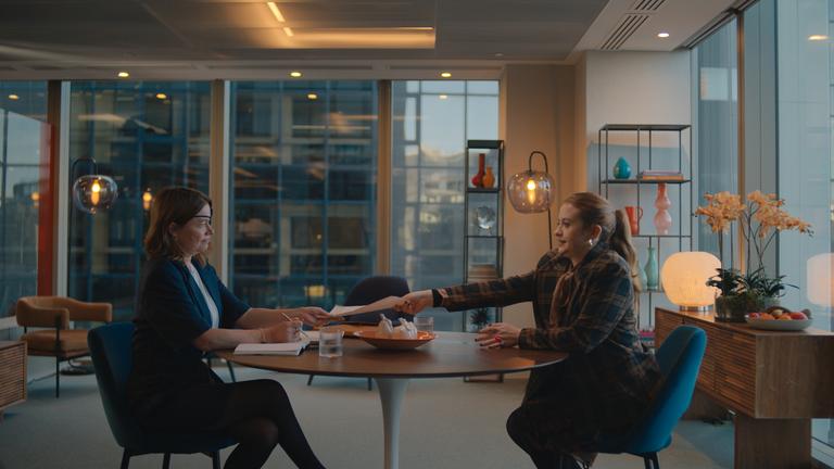 "Extraordinary": Colette (Robin Weaver) sitzt an einem runden Tisch in einem modernen, hellen Büro und nimmt ein Blatt Papier entgegen, das ihr Jen (Máiréad Tyers), die ihr gegenüber sitzt, überreicht. Mit ernster Miene blickt Colette dabei zu Jen.