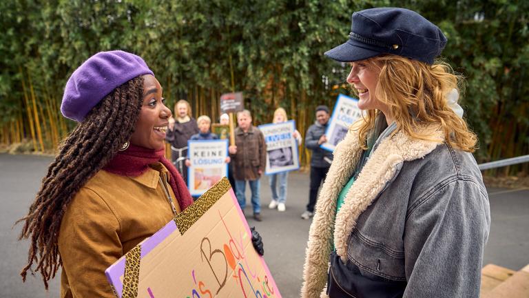 Nikita (Karmela Shako) und Lena (Anna Schimrigk) stehen sich im Freien gegenüber und strahlen sich gegenseitig an. Nikita hält ein Schild in der Hand mit der Aufschrift: "Lena's Body, Lena's Choice". Im Hintergrund sind Demonstranten mit Plakaten.