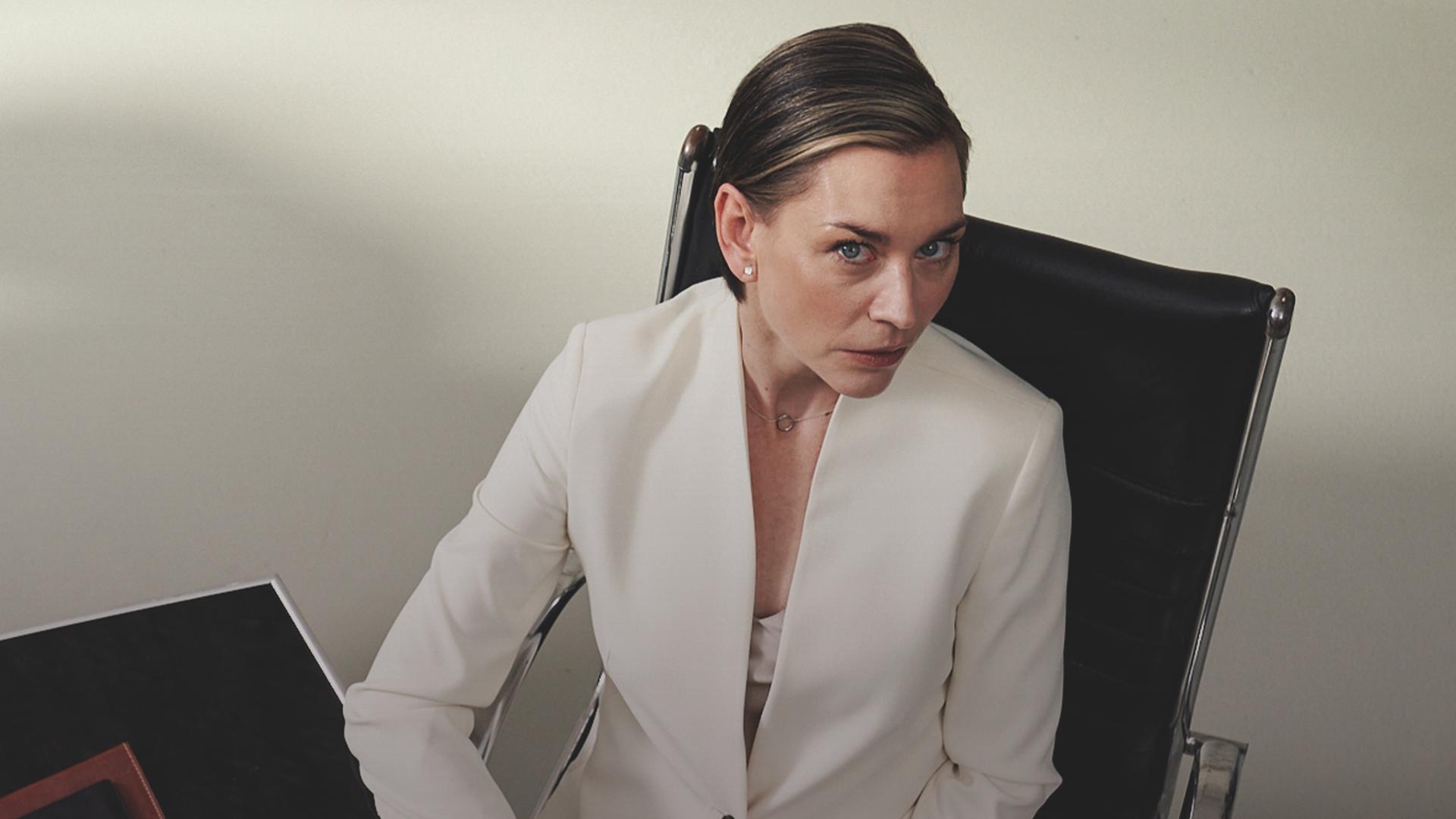 Juliane Ericksen (Christiane Paul), eine Frau mit kurzen glatten Haaren und in einem weißen Hosenanzug, sitzt an einem Schreibtisch und blick nach oben zur Kamera, die sie zu beobachten scheint.