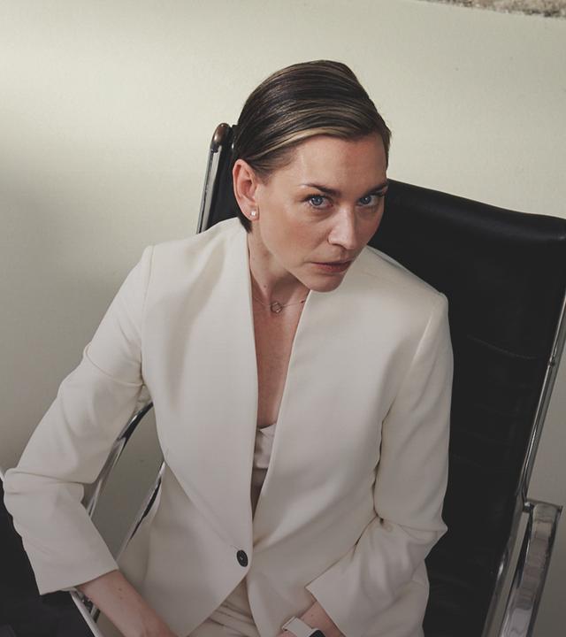 Juliane Ericksen (Christiane Paul), eine Frau mit kurzen glatten Haaren und in einem weißen Hosenanzug, sitzt an einem Schreibtisch und blick nach oben zur Kamera, die sie zu beobachten scheint.