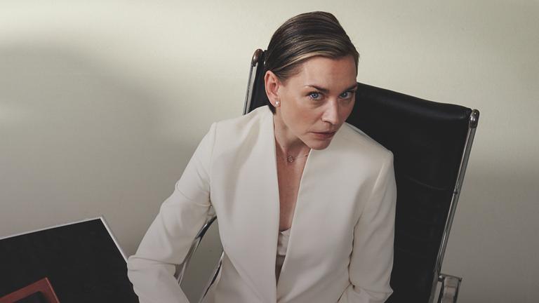 Juliane Ericksen (Christiane Paul), eine Frau mit kurzen glatten Haaren und in einem weißen Hosenanzug, sitzt an einem Schreibtisch und blick nach oben zur Kamera, die sie zu beobachten scheint.