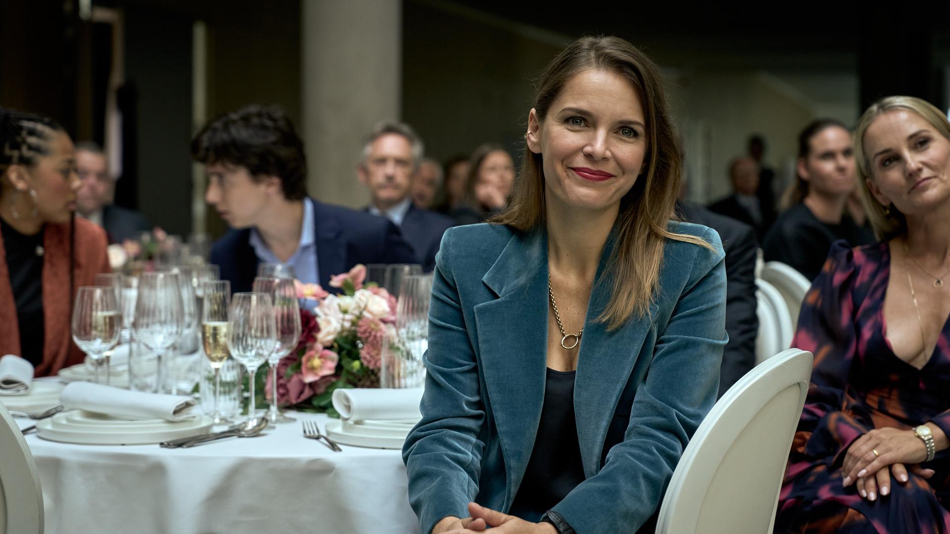 Isabelle (Hanna Verboom) sitzt an einem festlich gedeckten Tisch. Sie blickt nach vorne und lächelt. Hinter ihr sind Lola (J-Leeta Tijmes), Art (Roman Derwig), Marie (Peggy Vrijens) und weitere Gäste des Firmenempfangs zu sehen.