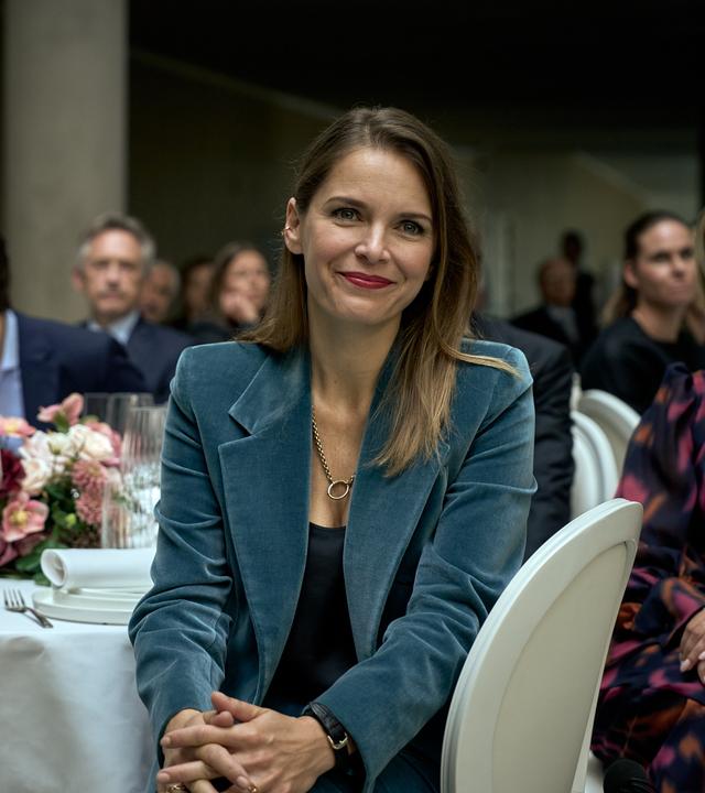 Isabelle (Hanna Verboom) sitzt an einem festlich gedeckten Tisch. Sie blickt nach vorne und lächelt. Hinter ihr sind Lola (J-Leeta Tijmes), Art (Roman Derwig), Marie (Peggy Vrijens) und weitere Gäste des Firmenempfangs zu sehen.