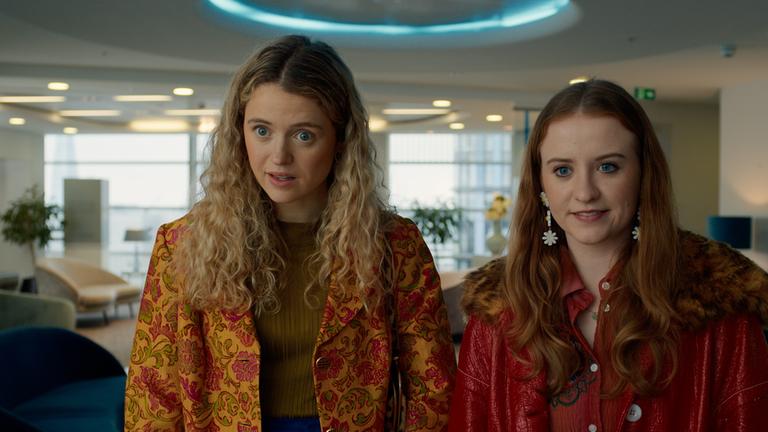 Jen (Máiréad Tyers, r.) und Carrie (Sofia Oxenham, l.) stehen nebeneinander in einem hellen, modern eingerichteten Raum mit Sesseln und großen Fenstern. Beide tragen farbenfrohe Outfits und blicken aufmerksam und leicht fragend geradeaus.