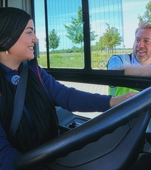 Eine junge Frau mit Kopftuch sitzt am Steuer eines Linienbusses. Neben dem offenen Fahrerfenster erklärt der Fahrlehrer wichtige Details zum Fahrzeug, während er in Richtung Außenspiegel zeigt. Die Szene zeigt eine praktische Fahrschulung, bei der der tote Winkel eines Busses erläutert wird. Die Szene zeigt eine praktische Fahrstunde.