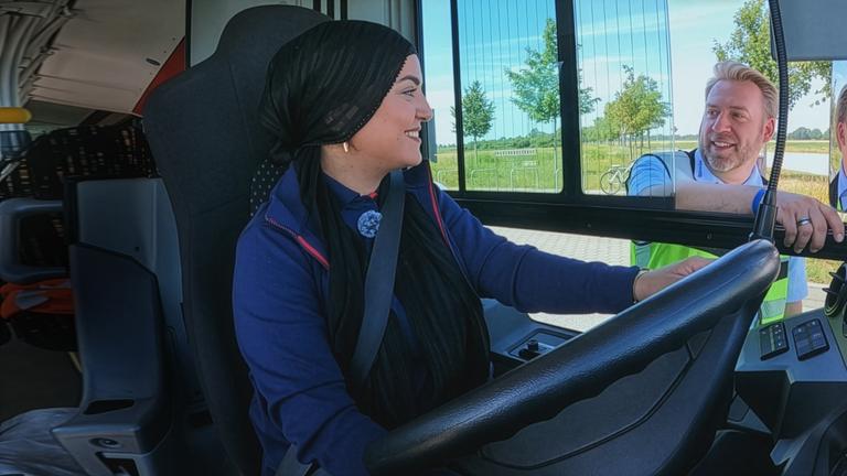 Eine junge Frau mit Kopftuch sitzt am Steuer eines Linienbusses. Neben dem offenen Fahrerfenster erklärt der Fahrlehrer wichtige Details zum Fahrzeug, während er in Richtung Außenspiegel zeigt. Die Szene zeigt eine praktische Fahrschulung, bei der der tote Winkel eines Busses erläutert wird. Die Szene zeigt eine praktische Fahrstunde.