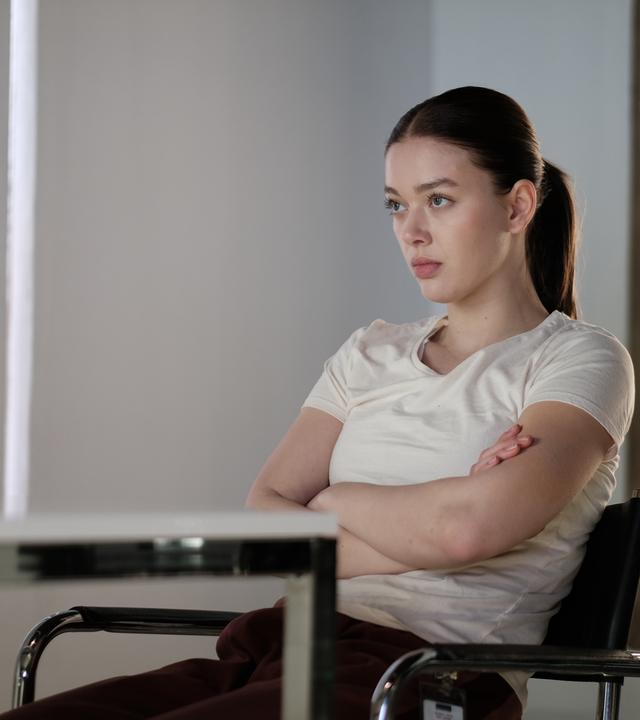 Innenraum mit weißen Wänden: Luce (Jessica Alexander) sitzt mit verschränkten Armen an einem Tisch und blickt konzentriert geradeaus. Im Hintergrund ist ein großes, bogenförmiges Fenster zu sehen.