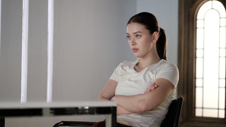 Innenraum mit weißen Wänden: Luce (Jessica Alexander) sitzt mit verschränkten Armen an einem Tisch und blickt konzentriert geradeaus. Im Hintergrund ist ein großes, bogenförmiges Fenster zu sehen.