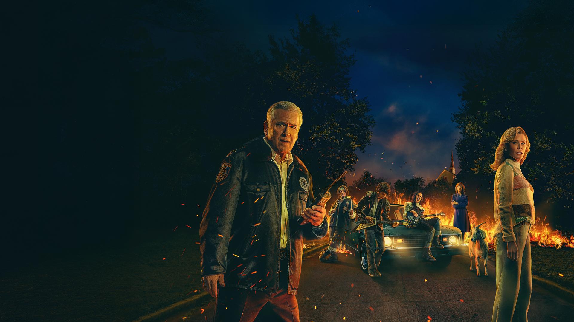 Im Vordergrund stehen ein Mann und eine Frau, die entsetzt in Richtung Kamera schauen. Im Hintergrund ist ein brennendes Auto zu sehen. Vor dem Auto sind zwei Jungs und ein Mädchen mit ihren Instrumenten zu sehen.