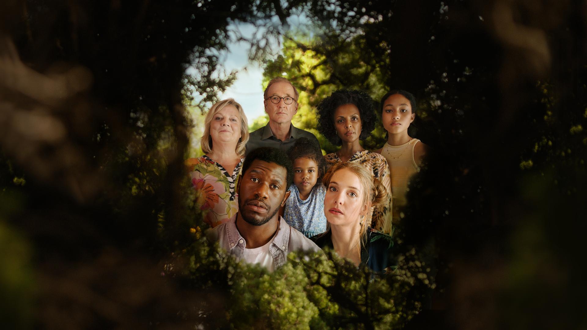I don't work here: In einer grünen Hecke ist ein Loch, durch das man einen Blick hinter die Hecke werfen kann. Dort ist eine Gruppe von Menschen zu sehen: Ganz vorne die Köpfe von Dawit (Akeem van Flodrop, 2.v.l.) und Laura (Sina Martens, 3.v.r.). Hinter ihnen stehen Heidi (Gabriela Maria Schmeide, l.), Walter (Peter Lohmeyer, 3.v.l.), Fiyori (Lousia Heinricht, M.), Lemlem (Dennenesch Zoudé, 2.v.r.) und Armana (Chioma Chukwuani, r.).