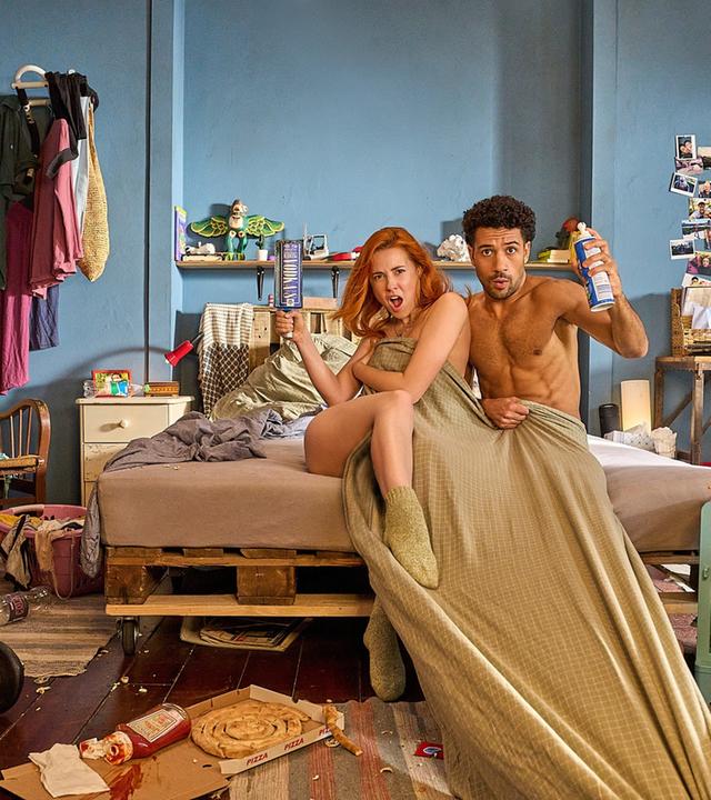 Caro (Meriel Hinsching) und Yannik (Rojan Juan Barani) sitzen in einem sehr unordentlichen Zimmer, halb zugedeckt, auf dem Bet und schauen frech nach vorne.