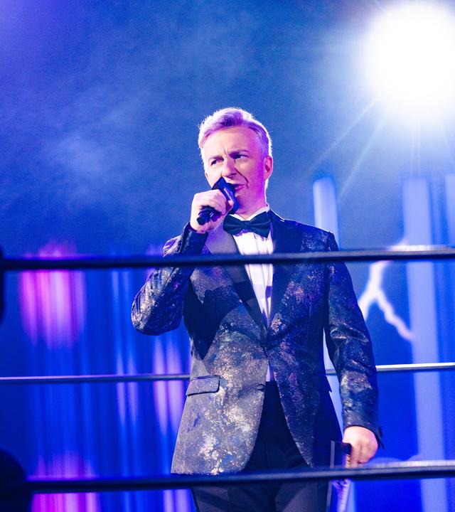 Jan Böhmermann ist als Ringrichter verkleidet. Er steht im Set der Sendung in einem Wrestlingring. Die Szene ist in Blautöne getaucht, ein Scheinwerfer strahlt hell von der Seite aus.