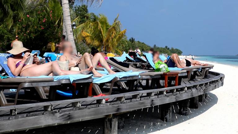 Touristen sonnen sich in nebeneinander aufgereihten Strandliegen. Im Hintergrund sind grüne Palmen zu sehen. Der Himmel ist wolkenlos blau und der Strand strahlend weiß.