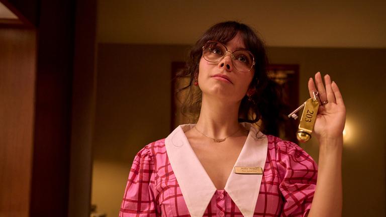 Millie steht hinter der Hotelrezeption und hält einen Schlüssel mit einem Anhänger hoch, auf dem "203" steht. Im Hintergrund sind ein Schrank mit Schlüsselboxen, ein Stuhl und eine Lampe sichtbar.