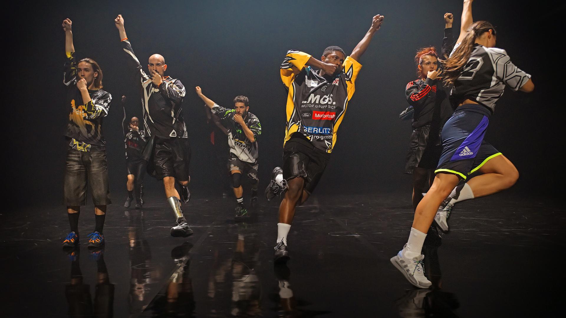 Eine Gruppe von acht Tänzerinnen und Tänzern in Motocross Shirts läuft Richtung Bühnenkante. Sie haben alle eine Faust in die Luftgestreckt und die andere zum Mund geführt. 