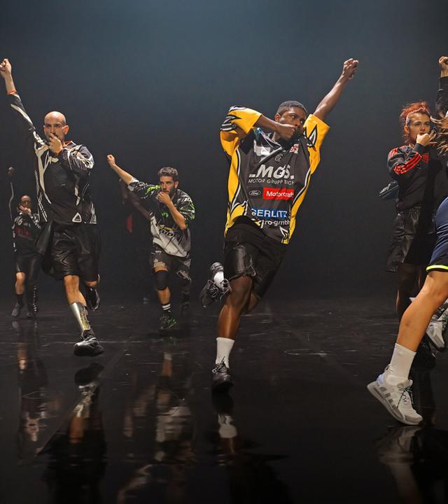 Eine Gruppe von acht Tänzerinnen und Tänzern in Motocross Shirts läuft Richtung Bühnenkante. Sie haben alle eine Faust in die Luftgestreckt und die andere zum Mund geführt. 