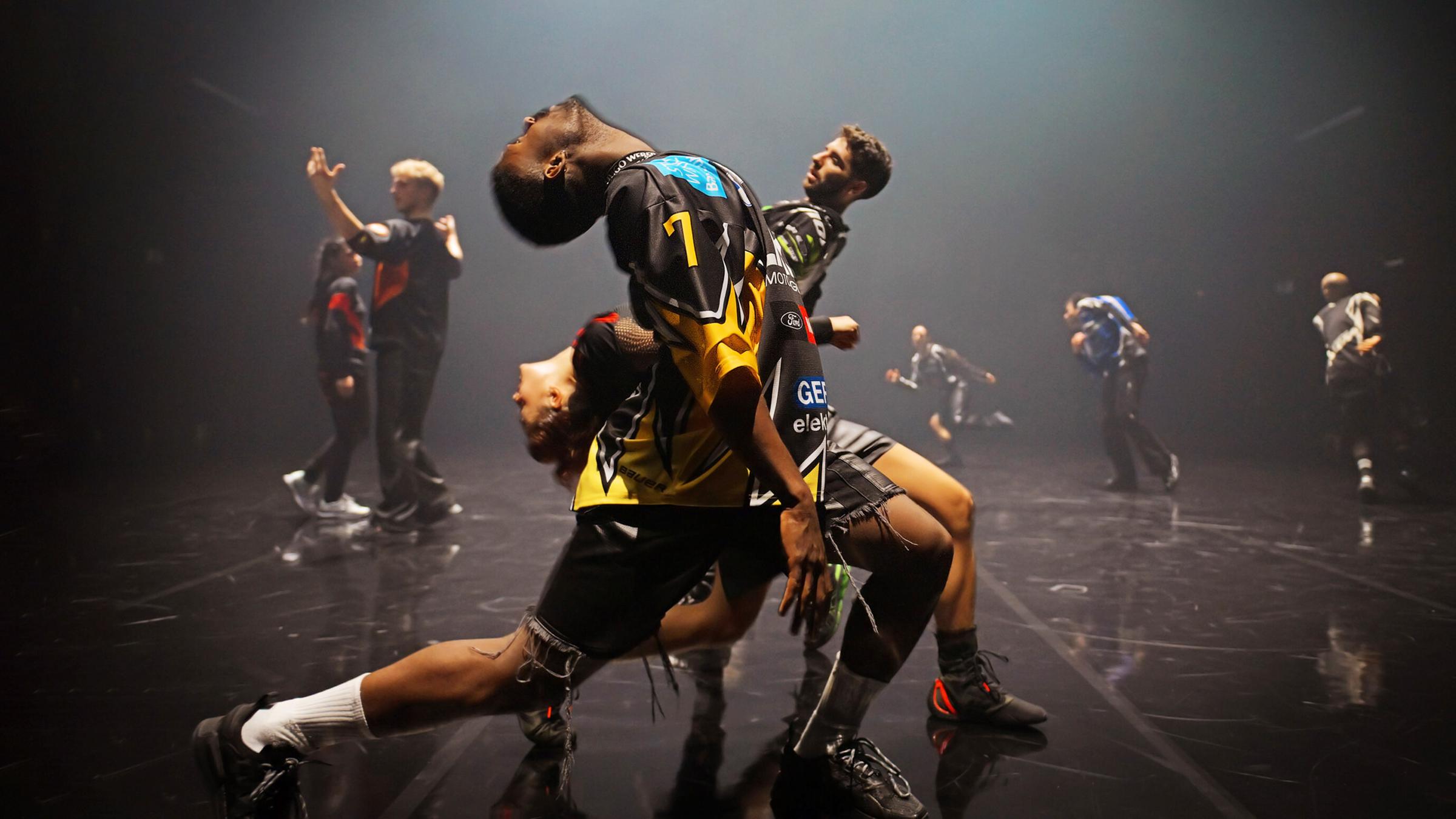 Zwei Tänzer und eine Tänzerin stehen seitlich zum Publikum hintereinander in einem weiten Ausfallschritt und beugen ihre Oberkörper nach hinten. Der vordere Tänzer und die Tänzerin nach links, der hinterste Tänzer nach rechts. Im Hintergrund sind fünf weitere Tänzerinnen und Tänzer zu erkennen.  