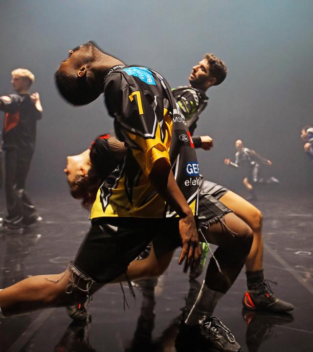 Zwei Tänzer und eine Tänzerin stehen seitlich zum Publikum hintereinander in einem weiten Ausfallschritt und beugen ihre Oberkörper nach hinten. Der vordere Tänzer und die Tänzerin nach links, der hinterste Tänzer nach rechts. Im Hintergrund sind fünf weitere Tänzerinnen und Tänzer zu erkennen.  