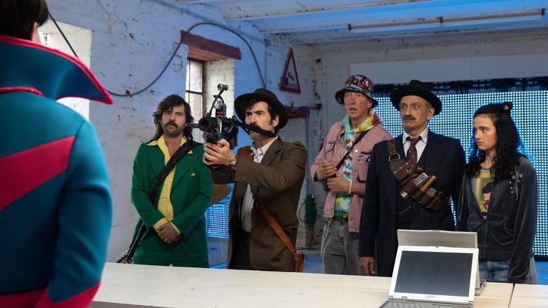 "Rembetis – die Geisterjäger – The Time Of My Life": Paris (Jasin Challah), Hektor (Samy Challah), Kevin (Martin Horn), Marcos (Pavlos Kourtidis) und Hekate (Sofia Koliofotos) stehen in einem kargen Raum mit blauer LED-Wand im Hintergrund einem Lotophagen gegenüber. Hektor hält eine pistolenartige Waffe auf den Lotophag gerichtet. Zwischen dem Lotophag und der Gruppe steht eine Holztheke auf der zwei Laptops platziert sind.