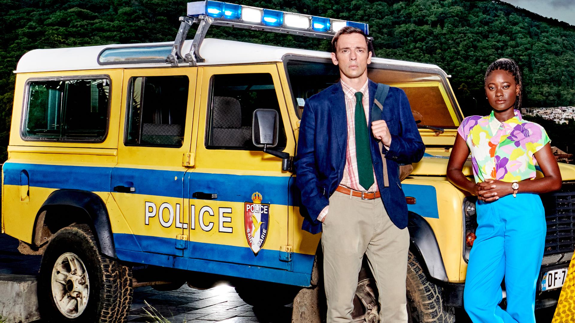 DI Neville Parker (Ralf Little) und Naomi Thomas (Shantol Jackson) lehnen an einem gelben Polizeiwagen mit blauen Streifen.