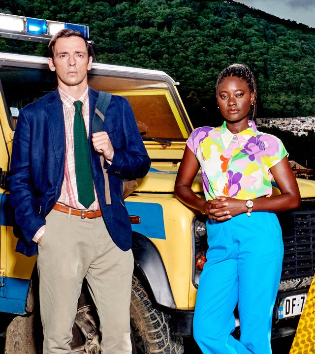 DI Neville Parker (Ralf Little) und Naomi Thomas (Shantol Jackson) lehnen an einem gelben Polizeiwagen mit blauen Streifen.