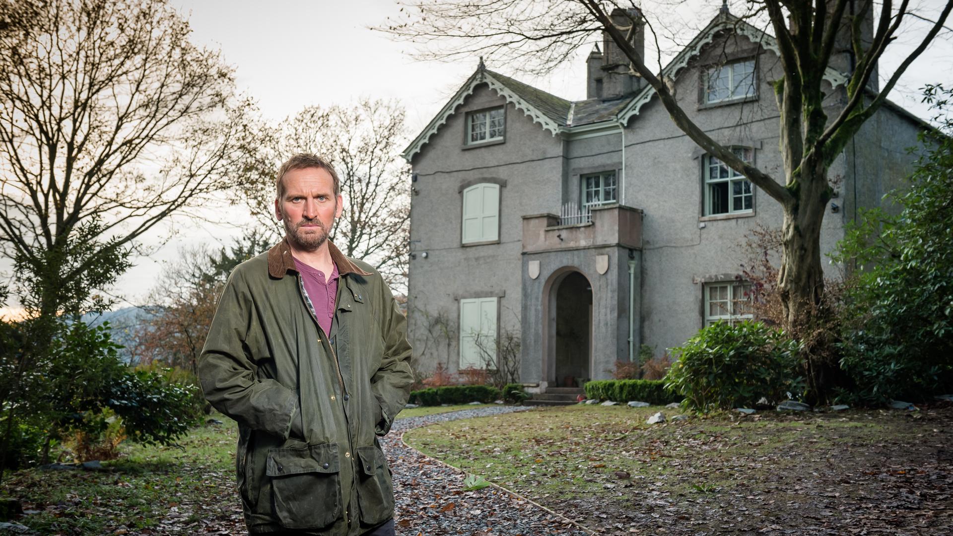 Robert Haleford steht mittig im Vorgarten vor seinem "Safe House". Er schaut geradeaus und hat beide Hände in seiner Jackentasche versteckt. Es ist herbstlich grau und sowohl rechts als auch links von ihm stehen vor dem Haus zwei Bäume, deren Laub im Vorgarten verteilt liegt.
