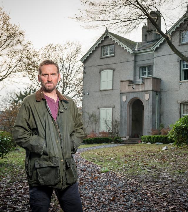 Robert Haleford steht mittig im Vorgarten vor seinem "Safe House". Er schaut geradeaus und hat beide Hände in seiner Jackentasche versteckt. Es ist herbstlich grau und sowohl rechts als auch links von ihm stehen vor dem Haus zwei Bäume, deren Laub im Vorgarten verteilt liegt.