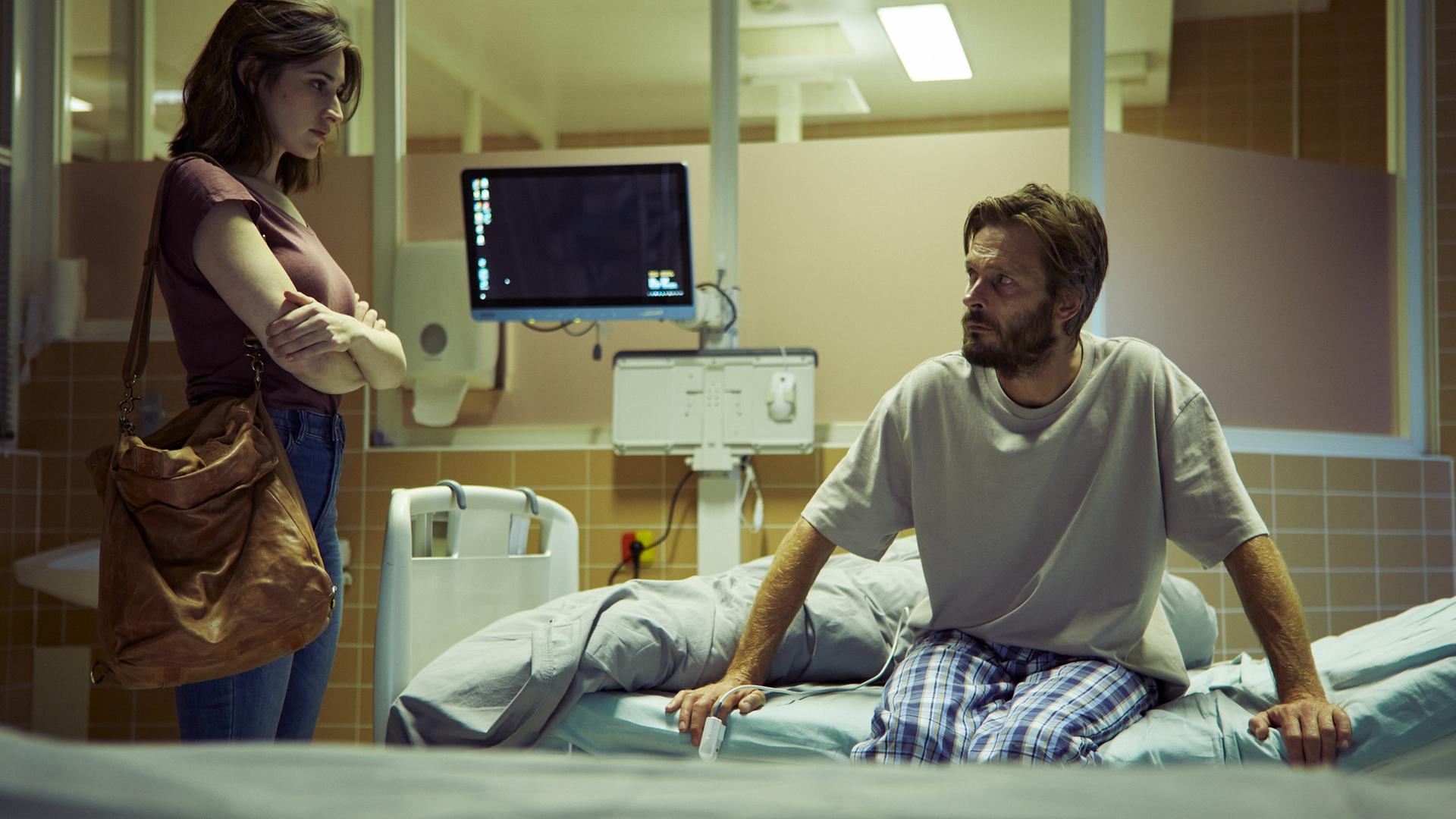 "Der Schatten - Schuld": Norah (Deleila Piasko) steht im Krankenhaus am Bett des Performance-Künstlers Wolfgang Balder (Andreas Pietschmann). Er sitzt am Rand des Bettes und stützt sich mit den Armen ab. Beide schauen sich an. Im Hintergrund ist ein Überwachungsmonitor zu sehen.