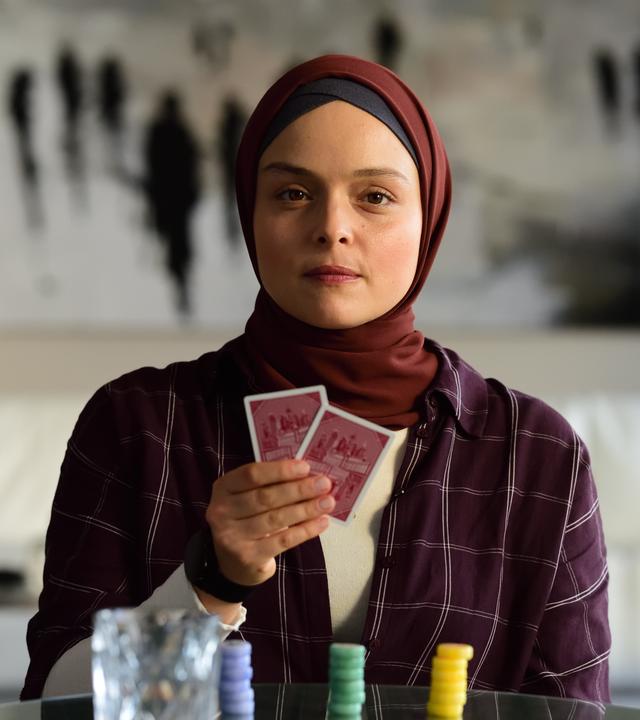 Ayla (Via Jikeli) sitzt in Vincents Wohnung am Tisch und hält zwei Karten vor sich.