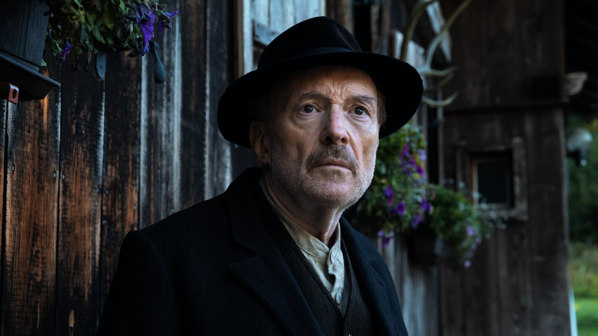 Sturm kommt auf: Schuster Kraus (Josef Hader), ein älterer Mann in Jacket und Hut, steht vor einem urigen Holzhaus. Sein Gesichtsausdruch ist besorgt.