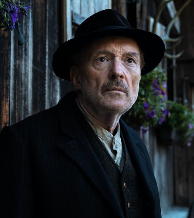 Sturm kommt auf: Schuster Kraus (Josef Hader), ein älterer Mann in Jacket und Hut, steht vor einem urigen Holzhaus. Sein Gesichtsausdruch ist besorgt.