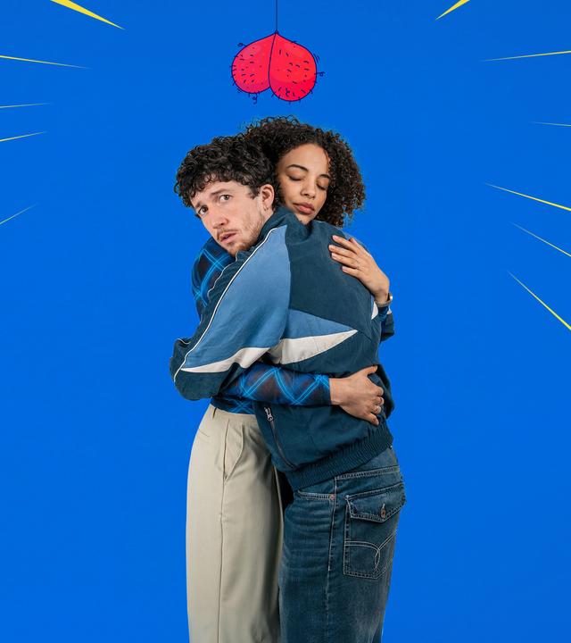 Noemi (Naomi Simmonds) und Julian (Maximilian Mundt) umarmen sich vor einem blauen Hintergrund. Julian schaut mit ängstlichem Blick in die Kamera, Noemi hat die Augen geschlossen. Über ihren Köpfen baumelt ein gemalter Hodensack.