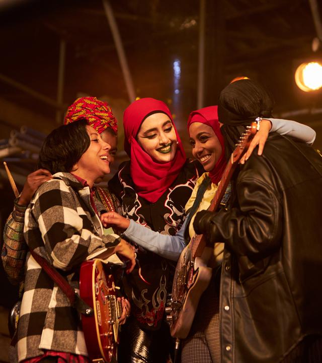 Saira (Sarah Kameela Impey), Bisma (Faith Omole), Ayesha (Juliette Motamed), Amina (Anjana Vasan) und Momtaz (Lucie Shorthouse) stehen eng umschlungen auf einer Bühne und lachen. Um sie herum leuchten warme Scheinwerfer. Saira und Amina haben jeweils eine E-Gitarre umgehängt.
