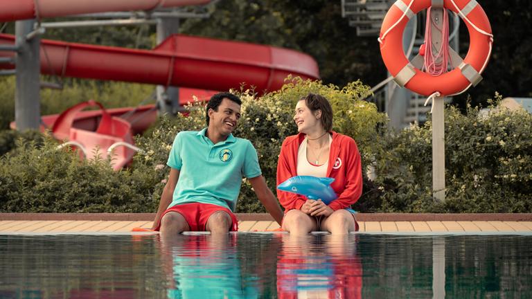 Mira (Anouk Elias) und Simon (Tyrell Otoo) sitzen am dem Becken des Freibads. Beide schauen sich an und lachen. Im Freibad sind keine weiteren Gäste zu sehen.
