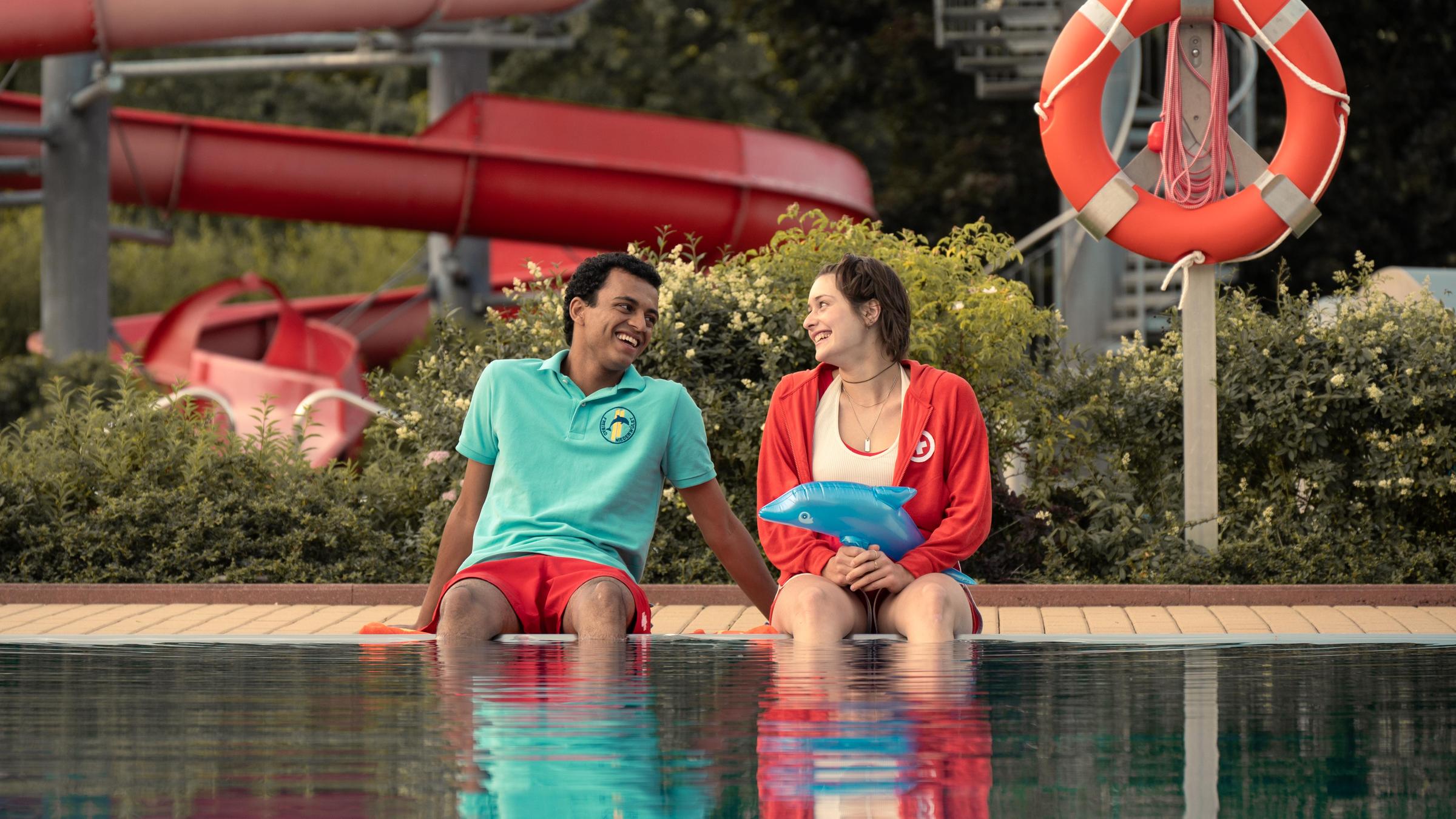 Mira (Anouk Elias) und Simon (Tyrell Otoo) sitzen am dem Becken des Freibads. Beide schauen sich an und lachen. Im Freibad sind keine weiteren Gäste zu sehen.