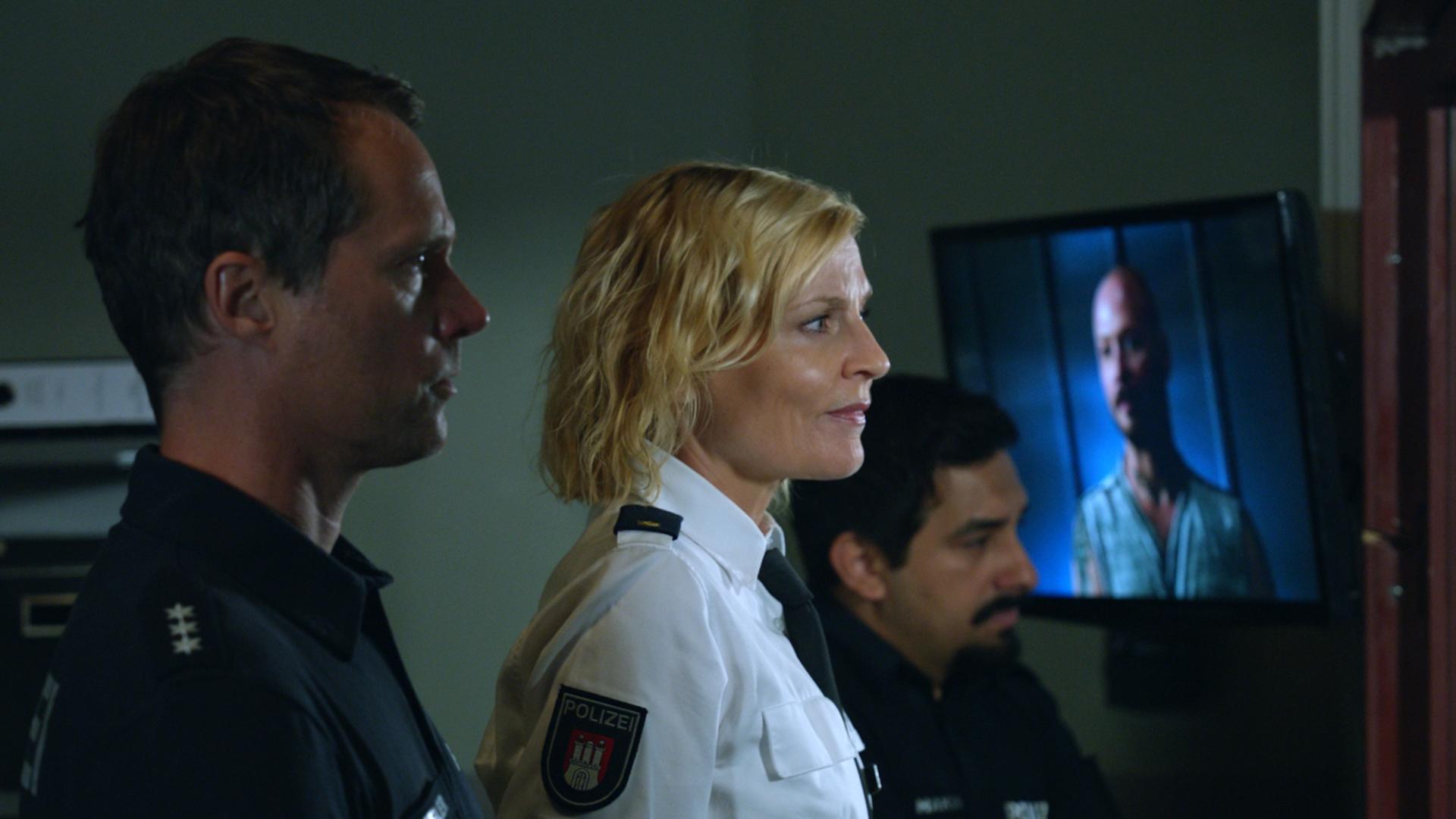 "Notruf Hafenkante - Schneckenhaus": Die Polizisten Mattes Seeler (Matthias Schloo) und Malik Hakim (Zejhun Demirov) stehen mit ihrer Kollegin Melanie Hansen (Sanna Englund) nebeneinander in einem Raum und beobachten durch ein Fenster eine Befragung, die im Raum daneben stattfindet.