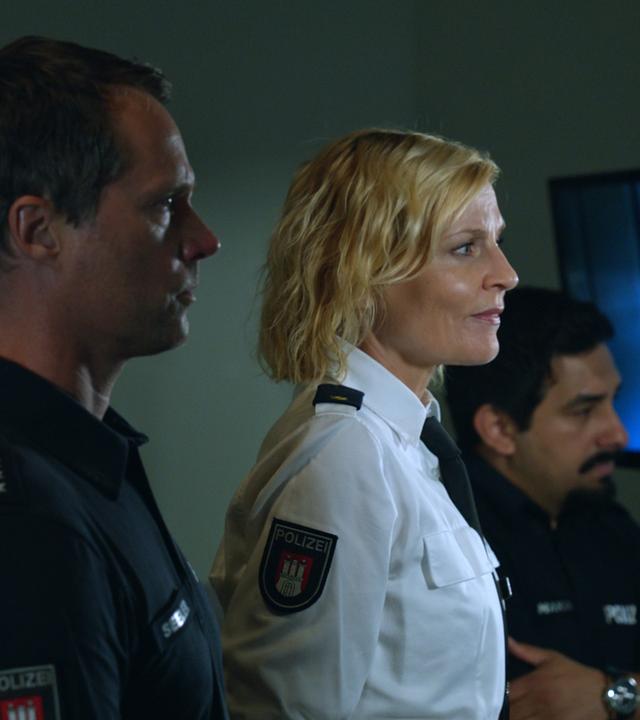 "Notruf Hafenkante - Schneckenhaus": Die Polizisten Mattes Seeler (Matthias Schloo) und Malik Hakim (Zejhun Demirov) stehen mit ihrer Kollegin Melanie Hansen (Sanna Englund) nebeneinander in einem Raum und beobachten durch ein Fenster eine Befragung, die im Raum daneben stattfindet.