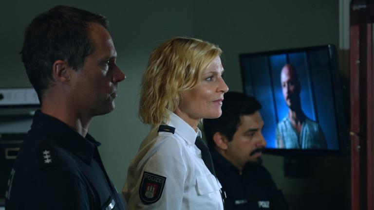"Notruf Hafenkante - Schneckenhaus": Die Polizisten Mattes Seeler (Matthias Schloo) und Malik Hakim (Zejhun Demirov) stehen mit ihrer Kollegin Melanie Hansen (Sanna Englund) nebeneinander in einem Raum und beobachten durch ein Fenster eine Befragung, die im Raum daneben stattfindet.
