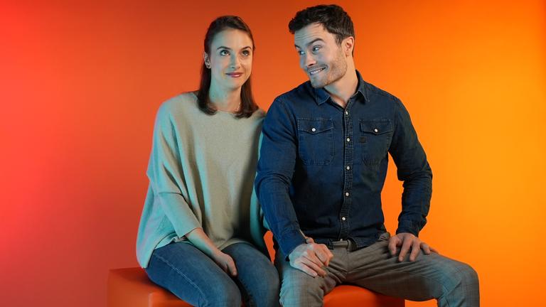 Ein Pärchen, eine Frau (links) und ein Mann (rechts) sitzen auf einem Hocker. Er schaut zu ihr rüber, sie blickt in die Luft.