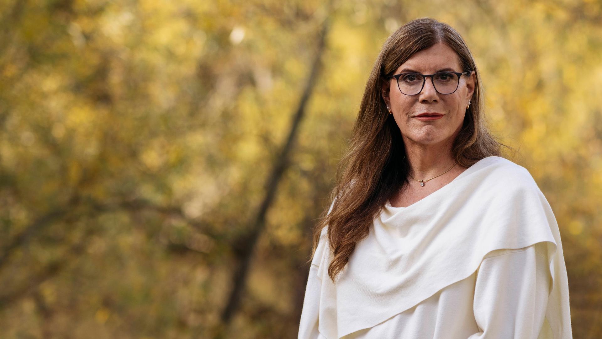 Constanze steht an eine kleine Mauer angelehnt in der Natur und lächelt in die Kamera. Im Hintergrund sind herbstlich anmutende Bäume zu sehen.