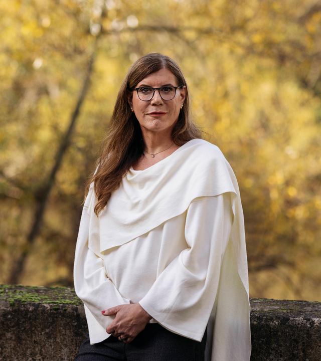 Constanze steht an eine kleine Mauer angelehnt in der Natur und lächelt in die Kamera. Im Hintergrund sind herbstlich anmutende Bäume zu sehen.