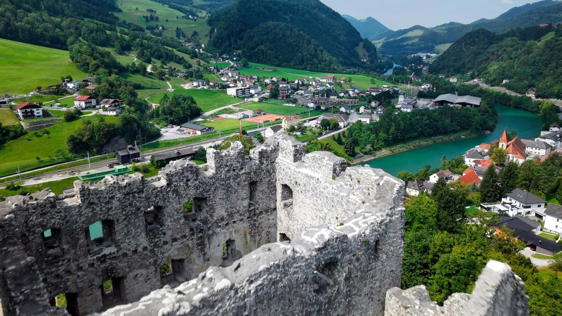 Das Bild zeigt die Ruine der Burg Losenstein, die auf einem Hang in einer malerischen Landschaft liegt. Die Burg hat massive, steinerne Mauern und einige Fensteröffnungen, die eine beeindruckende Aussicht bieten. Im Vordergrund ist der zerfallene Teil der Burg deutlich zu sehen, mit unregelmäßigen Kanten und vertrocknendem Mauerwerk.  Im Hintergrund erstreckt sich eine weite Hügellandschaft, die grün und bewaldet ist. Zwischen den Hügeln verlaufen ein Fluss und mehrere Siedlungen, die aus kleinen Häusern mit roten und weißen Dächern bestehen. Einige Straßen sind sichtbar, darunter eine Eisenbahnlinie, die entlang des Wassers verläuft.   Der Himmel ist teilweise bewölkt, und die Landschaft strahlt Ruhe und Naturverbundenheit aus. Die gesamte Szene vermittelt ein Gefühl von historischer Bedeutung und natürlicher Schönheit.