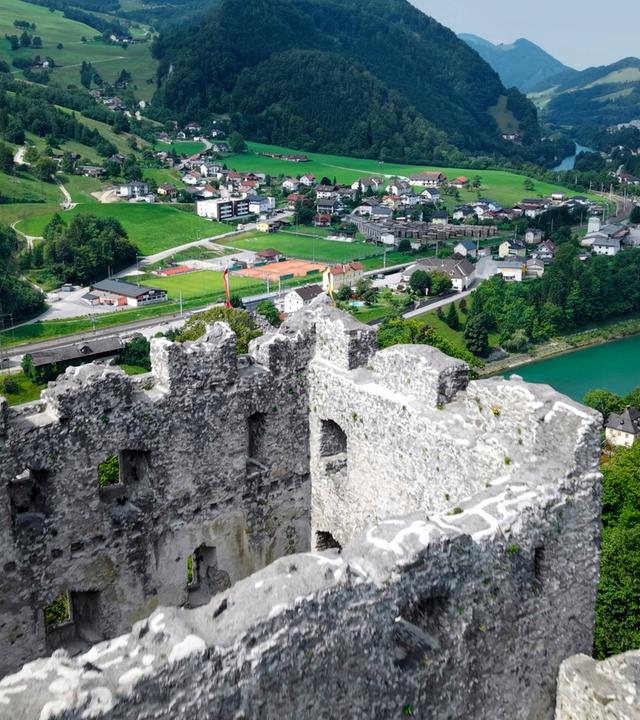 Das Bild zeigt die Ruine der Burg Losenstein, die auf einem Hang in einer malerischen Landschaft liegt. Die Burg hat massive, steinerne Mauern und einige Fensteröffnungen, die eine beeindruckende Aussicht bieten. Im Vordergrund ist der zerfallene Teil der Burg deutlich zu sehen, mit unregelmäßigen Kanten und vertrocknendem Mauerwerk.  Im Hintergrund erstreckt sich eine weite Hügellandschaft, die grün und bewaldet ist. Zwischen den Hügeln verlaufen ein Fluss und mehrere Siedlungen, die aus kleinen Häusern mit roten und weißen Dächern bestehen. Einige Straßen sind sichtbar, darunter eine Eisenbahnlinie, die entlang des Wassers verläuft.   Der Himmel ist teilweise bewölkt, und die Landschaft strahlt Ruhe und Naturverbundenheit aus. Die gesamte Szene vermittelt ein Gefühl von historischer Bedeutung und natürlicher Schönheit.