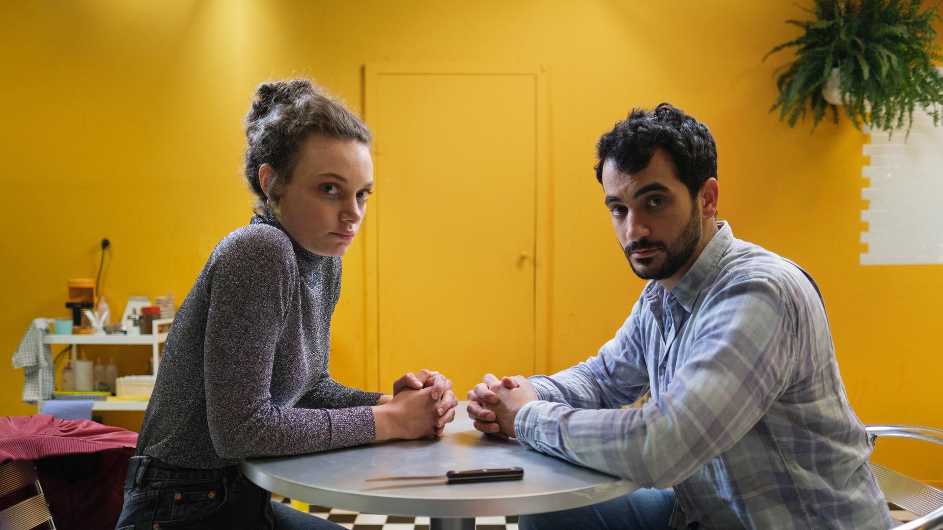 Nelly (Alina Heipe) sitzt mit einem bärtigen jungen Mann, Mervan (Timo Fakhravar) an einem kleinen runden Tisch. Auf dem Tisch liegt ein Küchenmesser. 