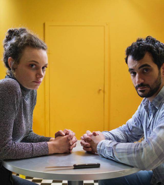 Nelly (Alina Heipe) sitzt mit einem bärtigen jungen Mann, Mervan (Timo Fakhravar) an einem kleinen runden Tisch. Auf dem Tisch liegt ein Küchenmesser. 